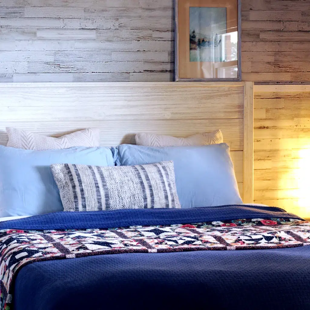 Quilt vs. Comforter: Understanding the Differences for Your Coziest Sleep 2 image displays a neatly made bed in a bedroom setting. The bed is adorned with a blue comforter and a variety of pillows in shades of blue and one with a cream and dark pattern. At the foot of the bed, there's a vibrant, multicolored quilt with intricate patterns.